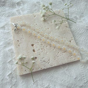 May include: A white and yellow beaded necklace with a silver clasp on a light beige textured surface.