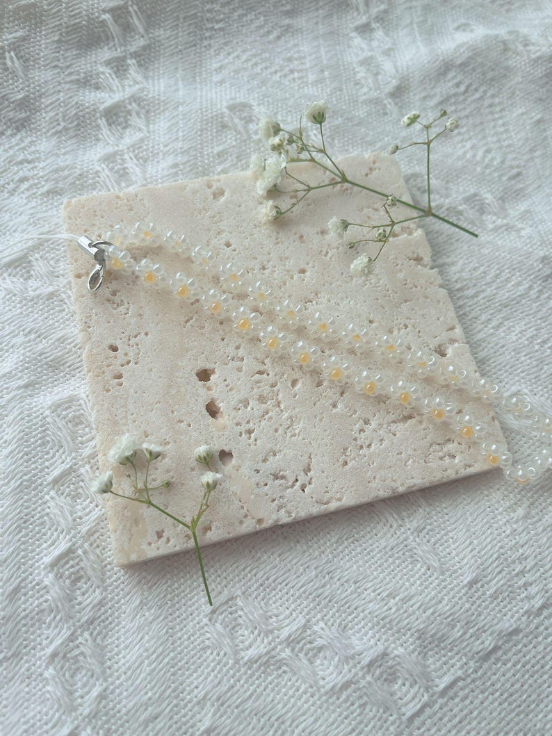 May include: A white and yellow beaded necklace with a silver clasp on a light beige textured surface.