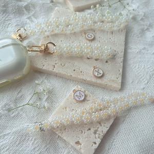 May include: Three white beaded phone straps with gold charms featuring the letters 'G', 'X', and 'M'. The charms are attached to the straps with a gold ring.