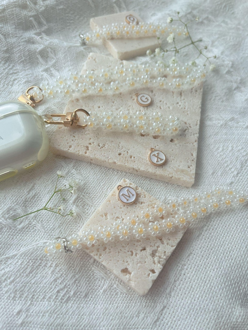 May include: Three white beaded phone straps with gold charms featuring the letters 'G', 'X', and 'M'. The charms are attached to the straps with a gold ring.