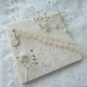 May include: A white beaded phone charm with a small gold charm with the letter 'G' on it. The charm is on a white marble surface with small white flowers.