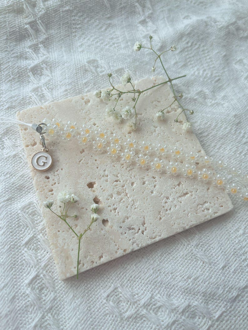 May include: A white beaded phone charm with a small gold charm with the letter 'G' on it. The charm is on a white marble surface with small white flowers.