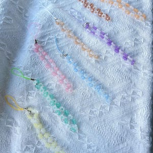 May include: A set of eight beaded phone charms, each featuring a different pastel colour and flower design. The charms are made with small, round beads and have a simple, delicate look.