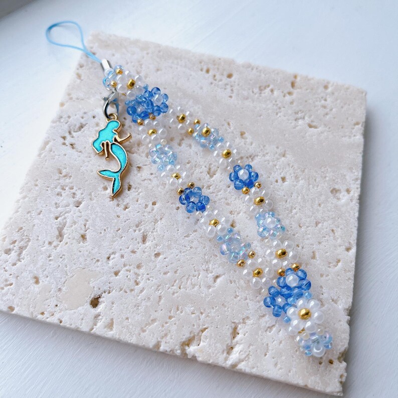 May include: A beaded phone charm with a mermaid charm. The beads are white, blue, and gold. The charm is turquoise blue.