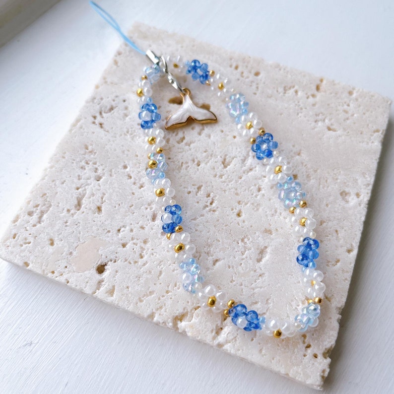 May include: A white and blue beaded phone charm with a gold accent and a white whale tail charm.