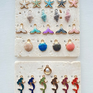 May include: Assortment of gold-tone charms with various sea-themed designs, including starfish, seashells, mermaids, and a pearl. The charms are arranged in rows on a white marble surface.
