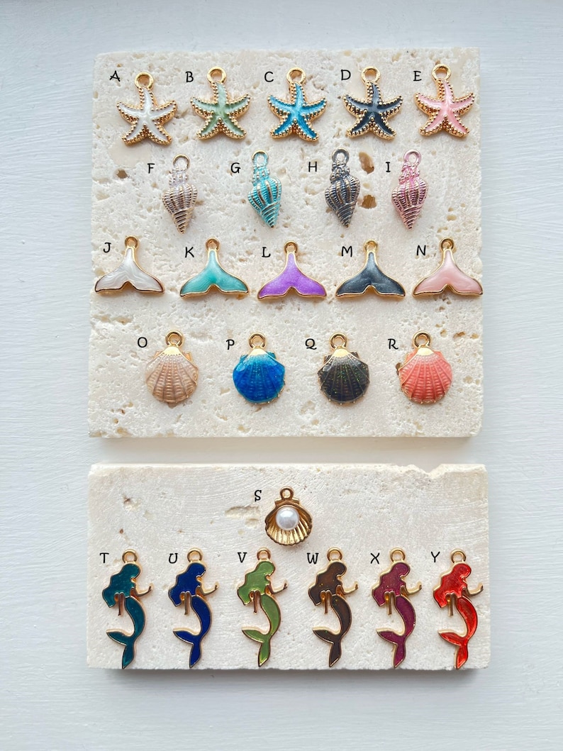 May include: Assortment of gold-tone charms with various sea-themed designs, including starfish, seashells, mermaids, and a pearl. The charms are arranged in rows on a white marble surface.
