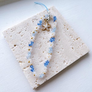May include: A white and blue beaded phone charm with a gold starfish charm. The charm is on a white marble surface.