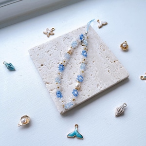 May include: A blue and white beaded phone charm with flower accents and a gold chain. The charm is on a white tile surface with other small decorative charms.