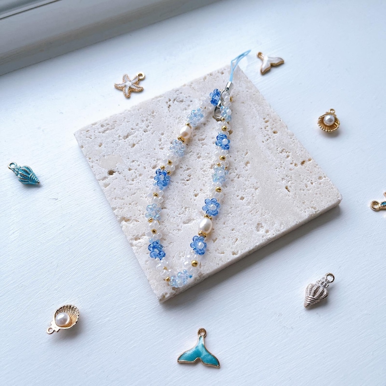 May include: A blue and white beaded phone charm with flower accents and a gold chain. The charm is on a white tile surface with other small decorative charms.