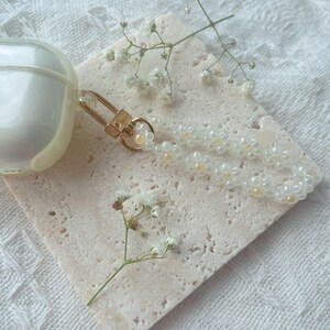 May include: A white and gold beaded phone charm with a gold clasp. The charm is on a white textured surface with small white flowers.