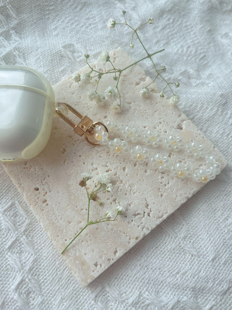 May include: A white and gold beaded phone charm with a gold clasp. The charm is on a white textured surface with small white flowers.