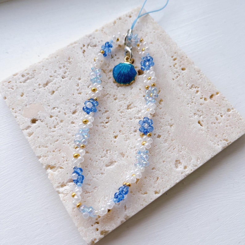 May include: A white and blue beaded phone charm with a small blue seashell charm. The beads are arranged in a gradient pattern, with white beads at the top and blue beads at the bottom.