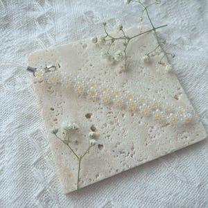 May include: A white beaded keychain with small yellow flower accents. The keychain is on a white textured surface with a small sprig of white flowers.