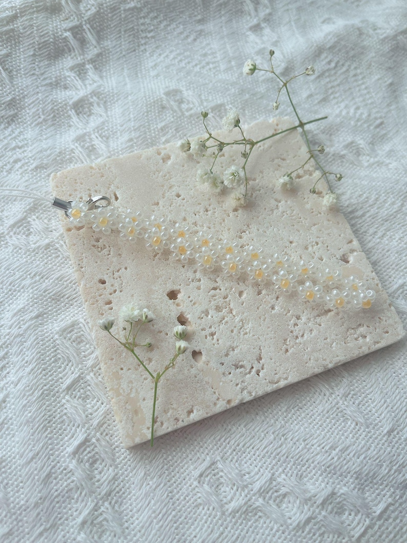 May include: A white beaded keychain with small yellow flower accents. The keychain is on a white textured surface with a small sprig of white flowers.