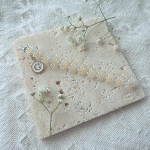 May include: A white and yellow beaded bracelet with a small charm featuring the letter "G" in white on a gold background.