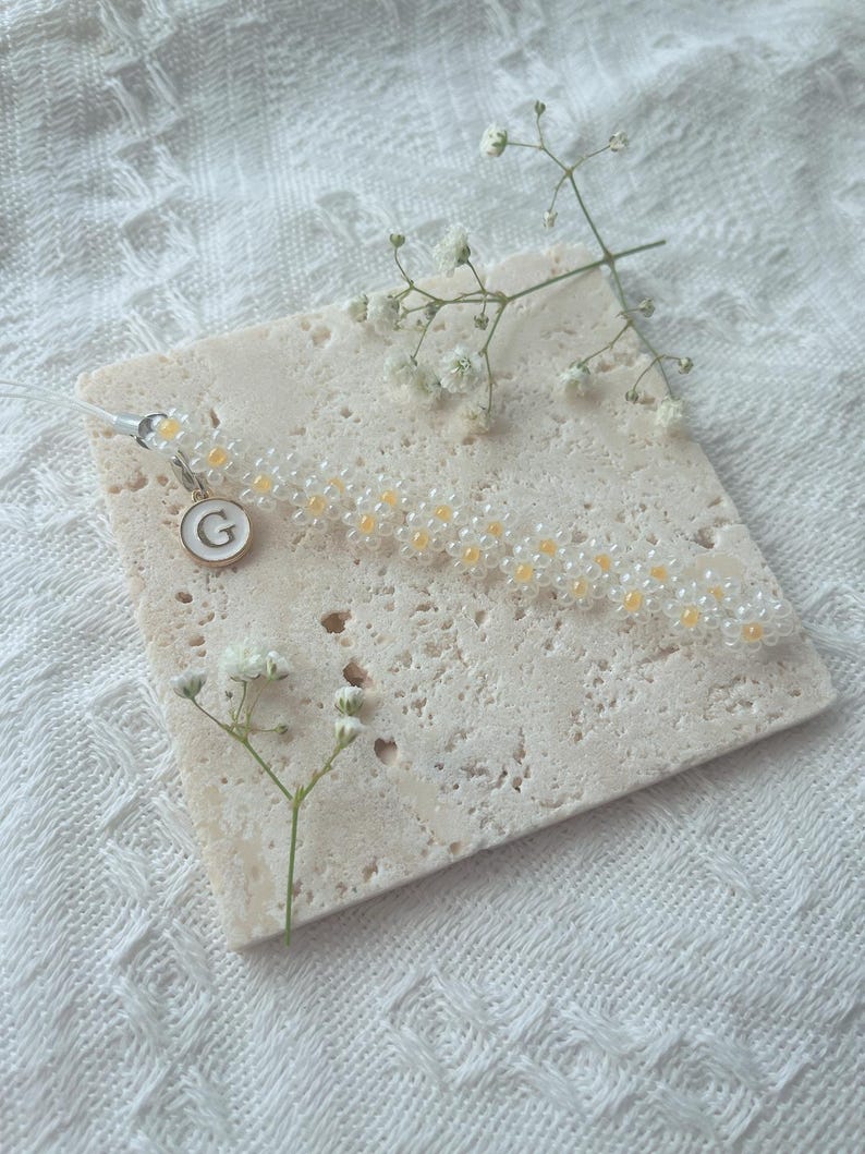 May include: A white and yellow beaded bracelet with a small charm featuring the letter "G" in white on a gold background.