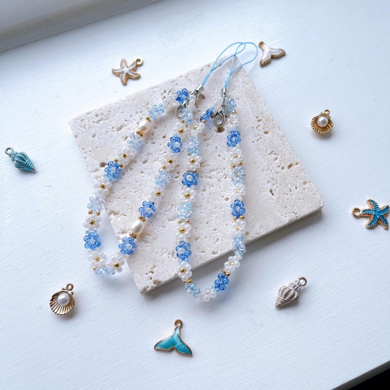 May include: Three beaded phone charms with blue and white flower beads and pearl accents. The charms are on a white marble surface with various blue and gold sea-themed charms.