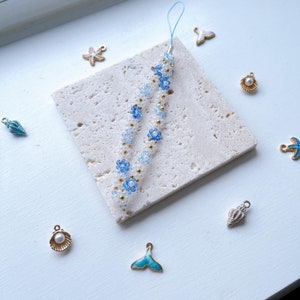 May include: A white and blue beaded phone charm with a floral design. The charm is on a white marble surface with other small charms, including a seashell, a starfish, and a mermaid tail.