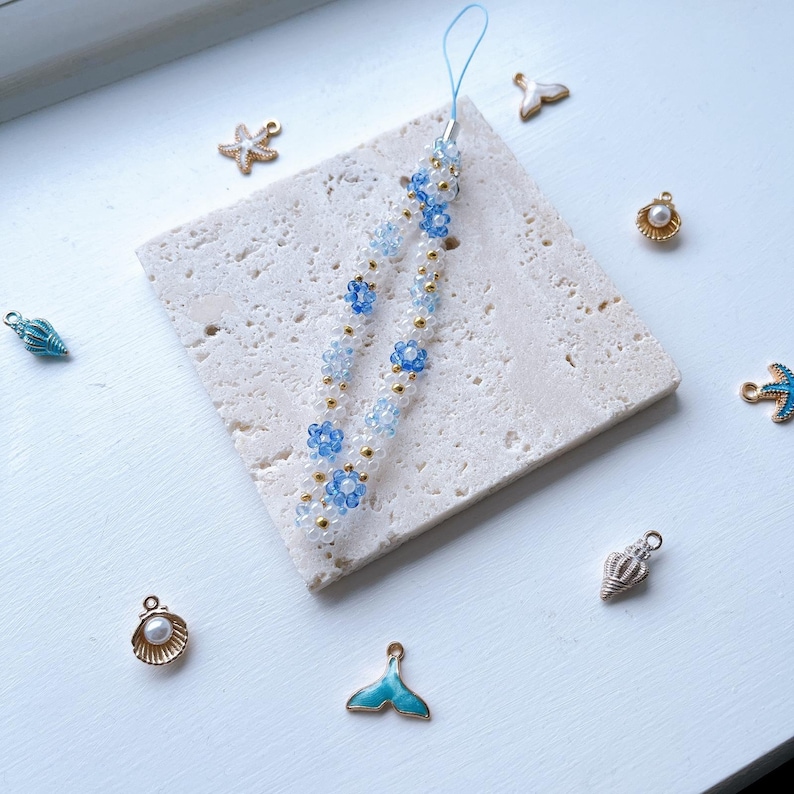 May include: A white and blue beaded phone charm with a floral design. The charm is on a white marble surface with other small charms, including a seashell, a starfish, and a mermaid tail.