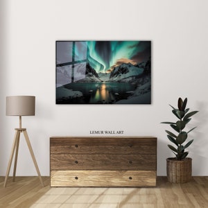 K&ouml;nnte beinhalten: Eine leuchtende gr&uuml;n-blaue Aurora Borealis spiegelt sich in einem stillen See wider, mit schneebedeckten Bergen im Hintergrund. Das Bild ist mit einem wei&szlig;en Rahmen versehen.