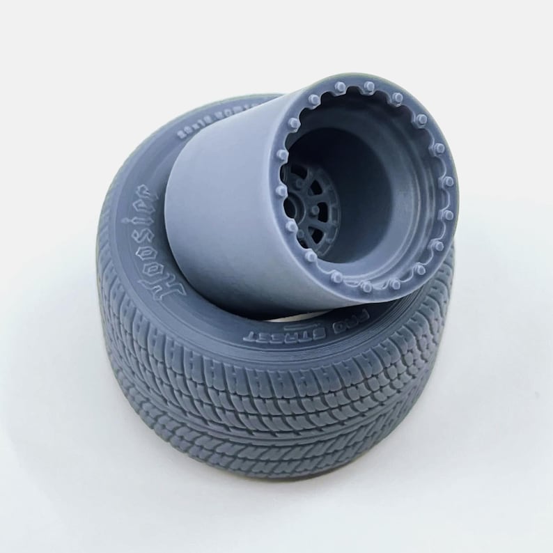 May include: A gray 3D printed model of a tire and wheel. The tire has a tread pattern and the words "Hoosier" and "Dragster" are visible on the sidewall. The wheel has a detailed design with a spoke pattern.