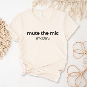 Puede incluir: Camiseta color crema con la frase "mute the mic" y el hashtag "#TODlife" impresos en negro. La camiseta está anudada en la parte inferior. Elementos decorativos incluyen un tapete tejido y un objeto floral.
