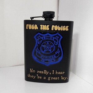 May include: Black metal flask with a gold inscription that reads "FUGK THE POLIGE" and a blue police badge with a gold star. The inscription below the badge reads "No really, I hear they're a great lay."
