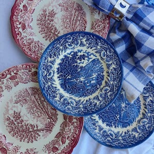 Könnte beinhalten: Eine Sammlung von Vintage-Keramikplatten mit Blumen- und Landschaftsdesigns. Zwei Teller haben ein rot-weißes Farbschema, während zwei andere ein blau-weißes Muster zeigen. Eine blau-weiß karierte Serviette wird von einem goldfarbenen Serviettenring gehalten.