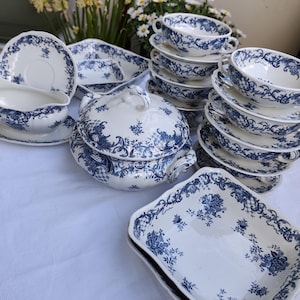 May include: A collection of white porcelain dishware with a blue floral pattern. The set includes soup bowls, saucers, serving dishes, and a covered tureen. The dishes are arranged on a white tablecloth.
