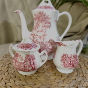 Könnte beinhalten: Ein weißes Keramik-Teeservice mit rotem Toile-Muster. Das Set beinhaltet eine Teekanne, eine Zuckerdose mit Deckel und einen Sahnekännchen. Das Muster zeigt eine ländliche Landschaft mit Bäumen und Gebäuden.
