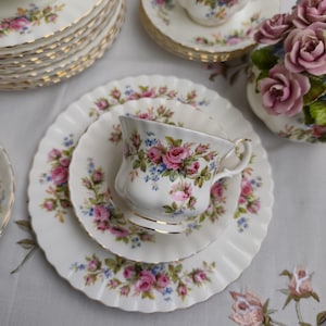 Könnte beinhalten: Weißes Porzellan-Teeservice mit rosa Rosen und blauen Blumenmotiven. Das Set umfasst Tassen, Untertassen und Teller mit Goldrand. Eine Vase mit rosa Rosen ergänzt das Set und sorgt für eine klassische, elegante Optik.