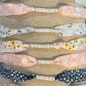 May include: A collection of fabric headbands with various floral patterns and beaded accents. Each headband features a different word spelled out in white letter beads, including "Love", "Peace", "Pray", "Grace", "Hope", "Joy", "Custom", and "XOXO".