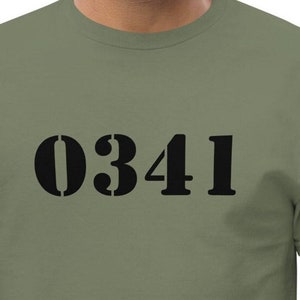 Könnte beinhalten: Olivgrünes T-Shirt mit der schwarzen Zahl "0341" auf der Vorderseite. Das Shirt ist einfarbig und hat kurze Ärmel. Die Person auf dem Bild trägt das T-Shirt.