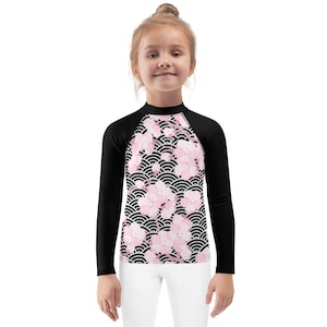 May include: A long-sleeved rash guard with a floral pattern. The shirt has black sleeves and a front panel with pink cherry blossoms on a black and white wave design. The shirt is worn by a young person.