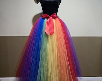 May include: A rainbow-colored tulle skirt displayed on a black mannequin. The skirt features vertical stripes of red, orange, yellow, green, blue, and purple. A red satin bow adorns the waist. The skirt is full and flowing.