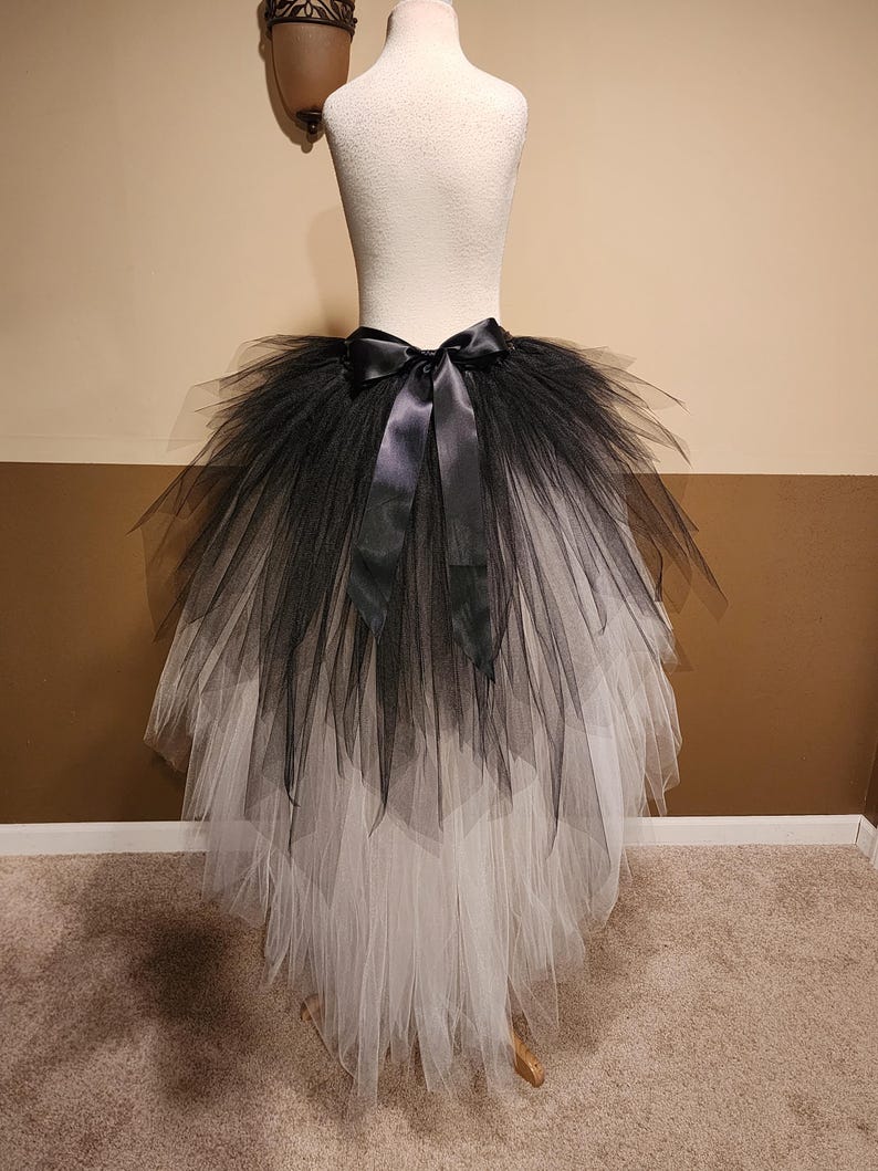 May include: A black and white tulle skirt with a satin bow. The skirt is displayed on a mannequin. The top layer is black, transitioning to white at the bottom. The skirt has a layered design.