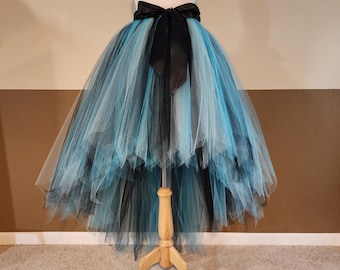 Pode incluir: Saia de tule em camadas em tons de preto, turquesa e azul claro, exibida em um manequim. A saia apresenta uma bainha assimétrica e um laço de cetim preto na cintura. Ideal para fantasias ou ocasiões especiais.