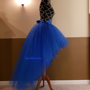 Puede incluir: Falda de tul azul real alta-baja con una faja de sat&eacute;n. La falda est&aacute; en un maniqu&iacute; y la cola de la falda est&aacute; arrastrando en el suelo. El texto "Sparkles tutu" es visible en la falda.