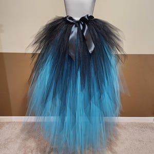 May include: A black and blue ombre tutu skirt with a satin ribbon bow at the waist. The skirt is made of layers of tulle, transitioning from black at the top to a vibrant blue at the bottom. The skirt is displayed on a mannequin.