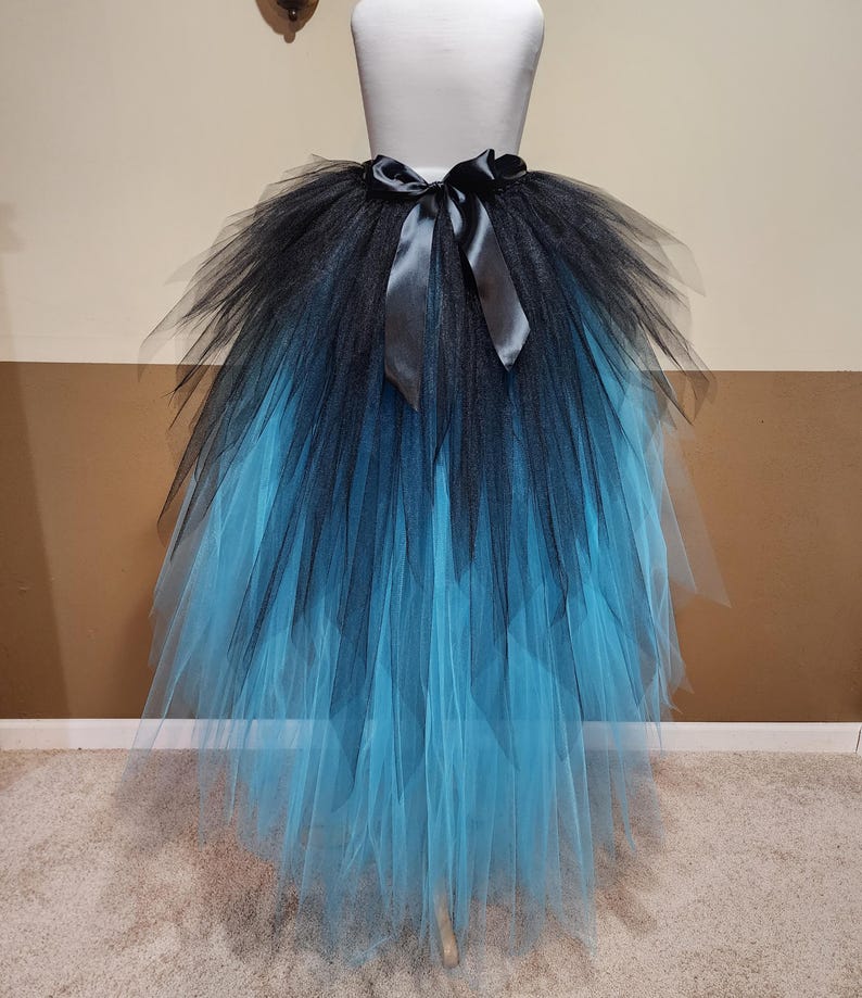 May include: A black and blue ombre tutu skirt with a satin ribbon bow at the waist. The skirt is made of layers of tulle, transitioning from black at the top to a vibrant blue at the bottom. The skirt is displayed on a mannequin.