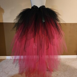 May include: A black and pink ombre tulle skirt displayed on a mannequin. The skirt features a black waistband with a black bow. The tulle transitions from black at the top to a vibrant pink at the bottom, creating a dramatic effect.