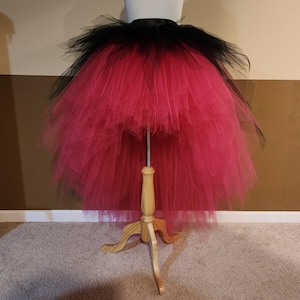 May include: A vibrant, layered tutu skirt in black and hot pink, displayed on a dress form. The skirt features a black waistband and a voluminous, high-low design. The tulle layers create a full, textured effect, perfect for costumes or dance.