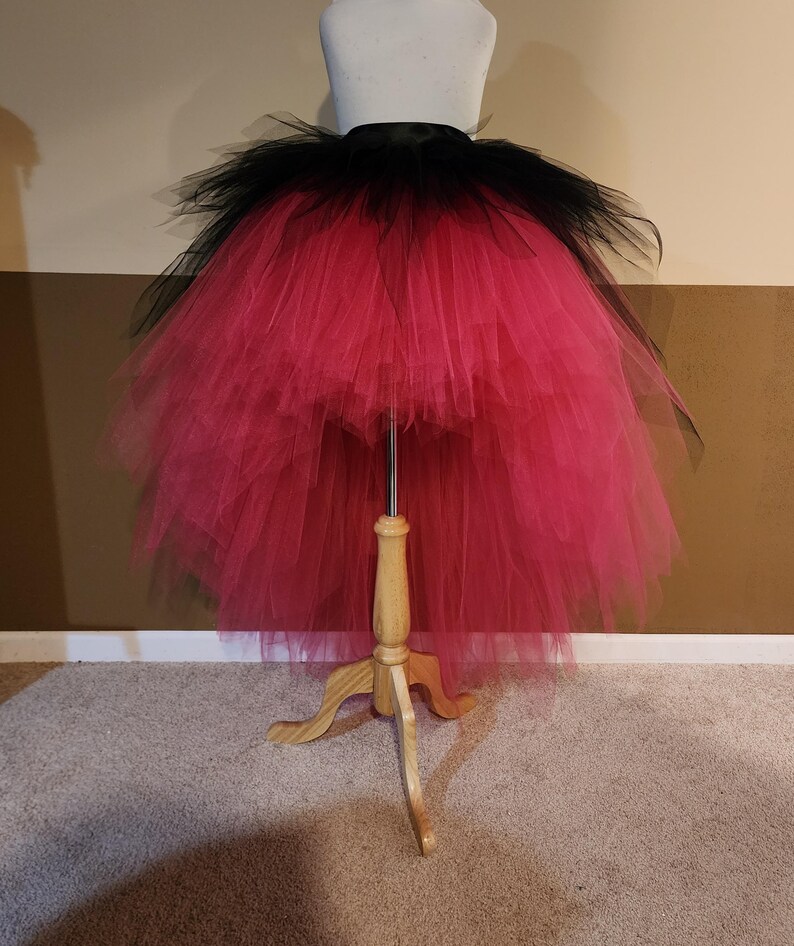 May include: A vibrant, layered tutu skirt in black and hot pink, displayed on a dress form. The skirt features a black waistband and a voluminous, high-low design. The tulle layers create a full, textured effect, perfect for costumes or dance.