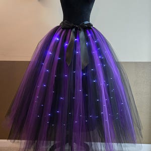 May include: A black tulle tutu skirt with alternating purple and black panels. The skirt is adorned with small, twinkling blue lights, and a black satin bow at the waist. The skirt is displayed on a black mannequin.