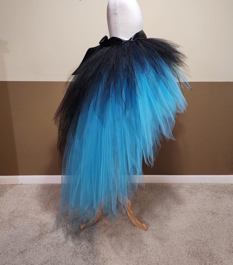 May include: A black and blue tutu skirt displayed on a mannequin. The skirt features a black waistband and a layered design that fades from black to a vibrant blue. The tulle skirt is designed to be asymmetrical, with a longer length in the back.