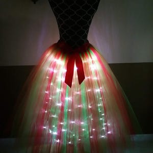 May include: A festive tutu skirt on a mannequin, illuminated with red, green, and white lights. The skirt features layers of tulle and a red bow at the waist. The lights create a glowing effect, perfect for a holiday or special occasion.