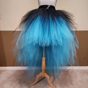 May include: A black and turquoise tutu skirt displayed on a dress form. The skirt features a black waistband and a layered design, transitioning from black at the top to turquoise at the bottom. The tulle skirt is full and flowing.
