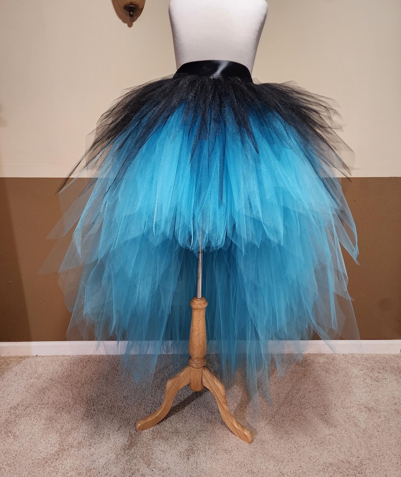 May include: A black and turquoise tutu skirt displayed on a dress form. The skirt features a black waistband and a layered design, transitioning from black at the top to turquoise at the bottom. The tulle skirt is full and flowing.