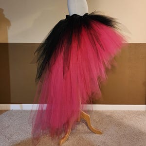 May include: A black and hot pink tulle skirt displayed on a mannequin. The skirt has a high-low design, with black tulle at the top that fades into a vibrant pink. The skirt is gathered at the waist with a black ribbon.